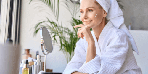 Woman in spa robe applying skincare, smiling at mirror with natural light. Beauty routine, self-care, skincare products nearby.