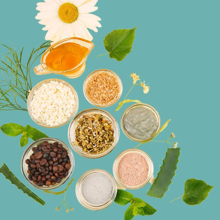 Natural skincare ingredients flat lay with herbs, clay, aloe, coffee, honey, and essential oils on a teal background.
