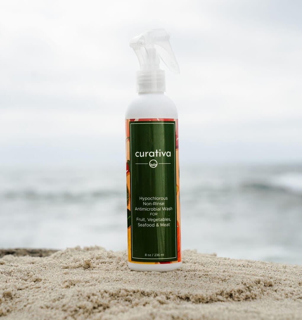 Curativa antimicrobial wash bottle on sandy beach, ocean background; non-rinse cleaner for fruits, vegetables, seafood, meat.