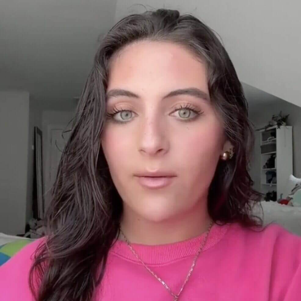 Portrait of a woman with long dark hair wearing a pink sweater in a bright room.