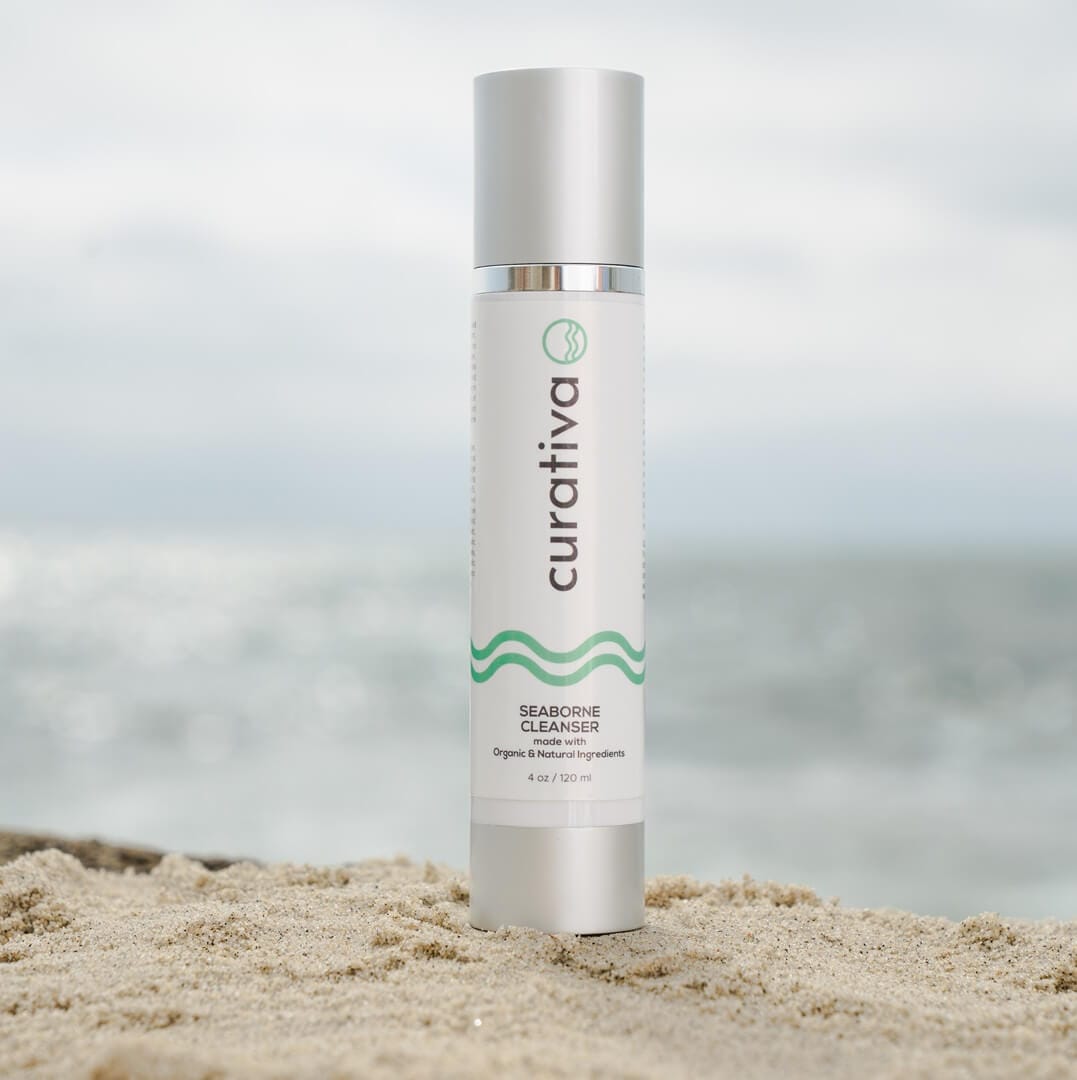 Curativa Seaborne Cleanser on sandy beach with ocean backdrop, organic skincare product, natural ingredients, 120ml bottle.