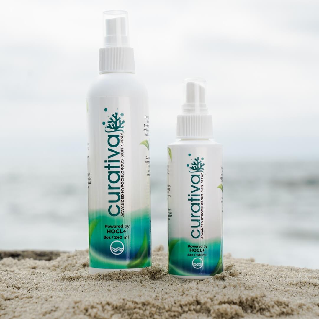 Curativa hypochlorous skin spray bottles on sandy beach, ocean background.