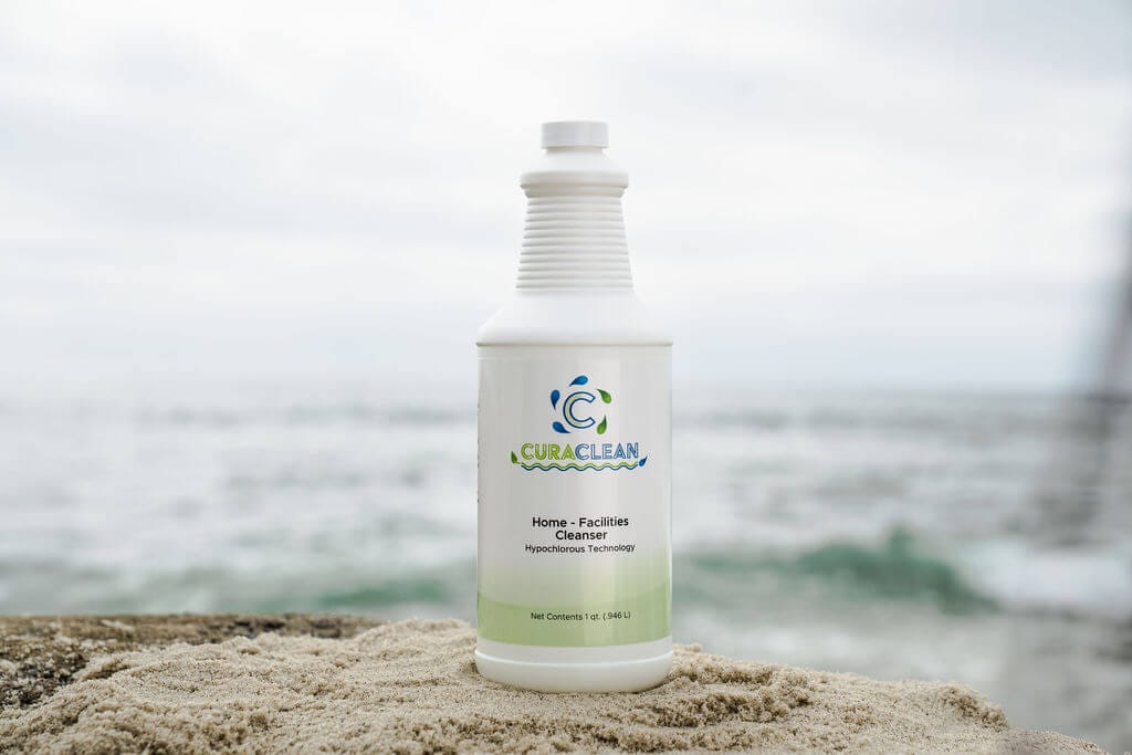 Curativa-64 home facilities cleanser bottle on sandy beach with ocean in background, showcasing eco-friendly cleaning product.