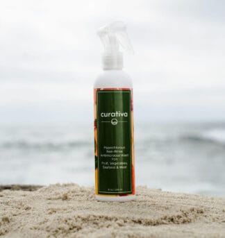Curativa antimicrobial wash bottle on sandy beach, ocean background; non-rinse cleaner for fruits, vegetables, seafood, meat.